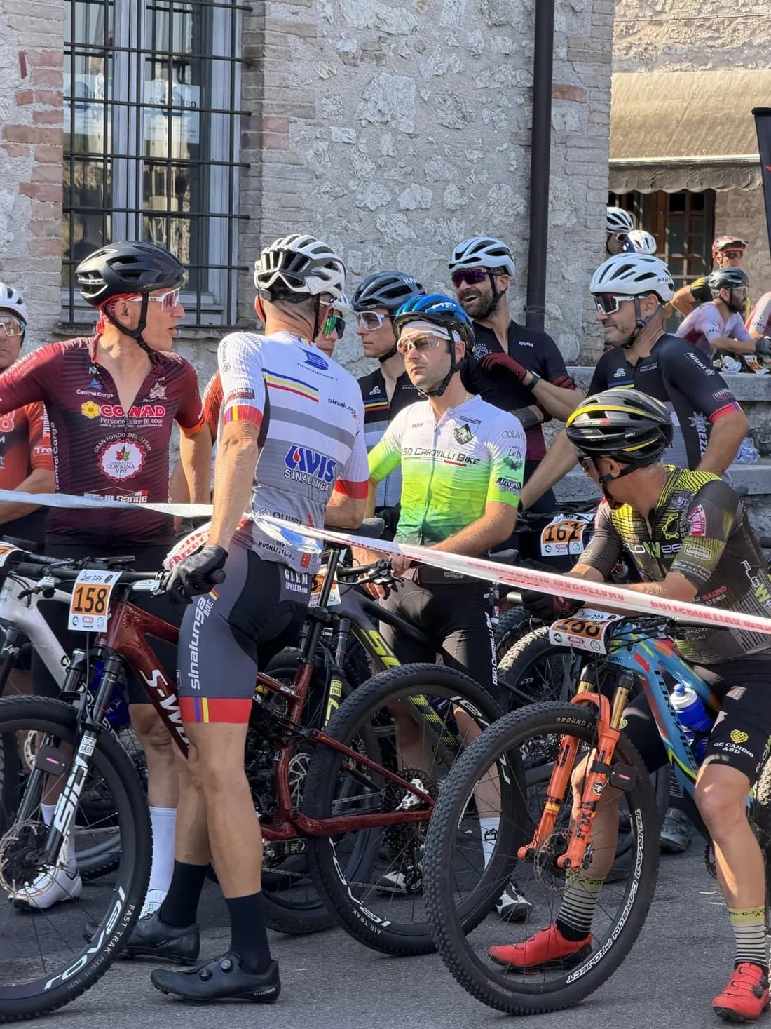 Atleti in attesa in griglia con le mountain bike prima della partenza, davanti al municipio di Arrone