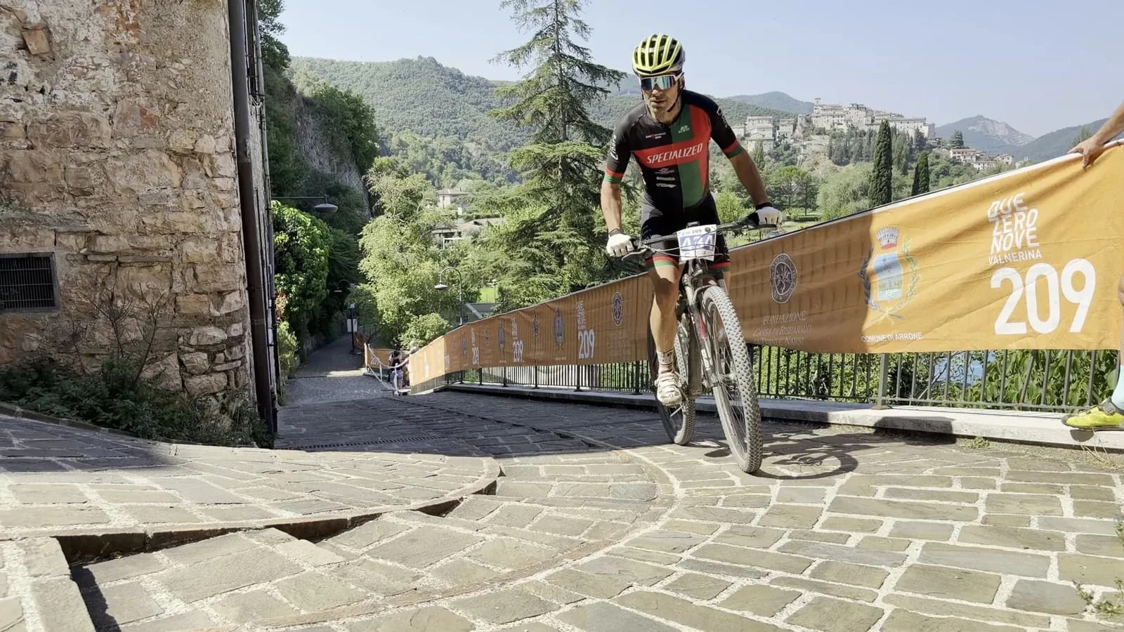 Atleta in maglia Specialized supera la rampa in pietra di Arrone lungo le transenne gialle della Marathon 209, il borgo e le colline della Valnerina sullo sfondo
