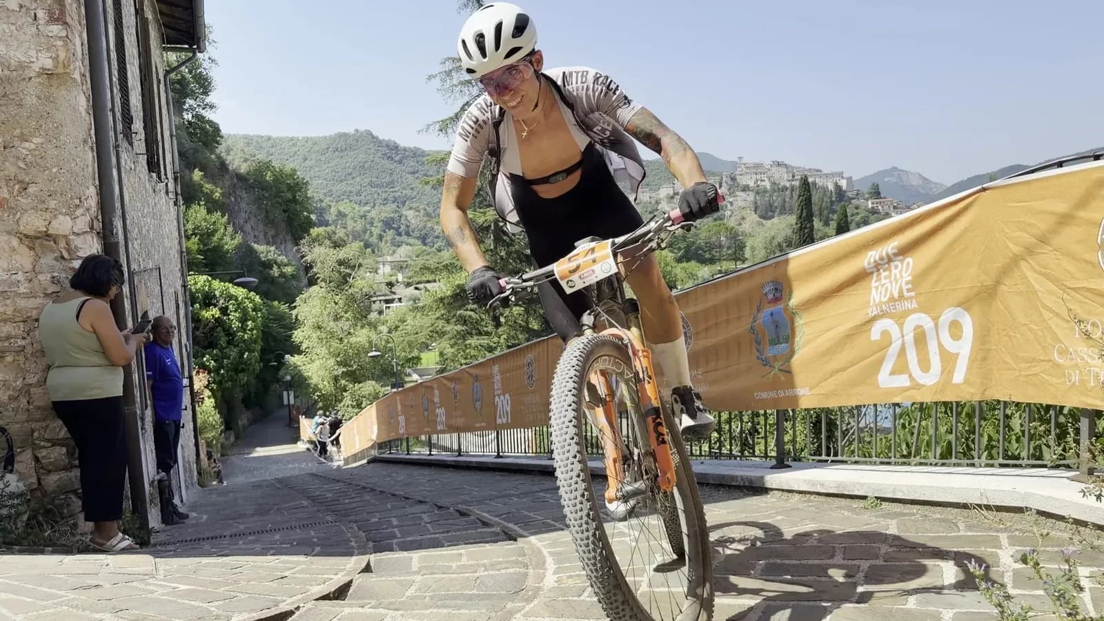 Atleta in mountain bike affronta in piedi sui pedali la rampa in pietra del borgo di Arrone, transenne della Marathon 209 e colline della Valnerina sullo sfondo