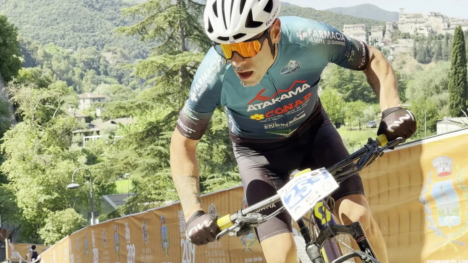 Primo piano di un atleta in piedi sui pedali nel tratto più ripido, con la vallata del Nera alle spalle