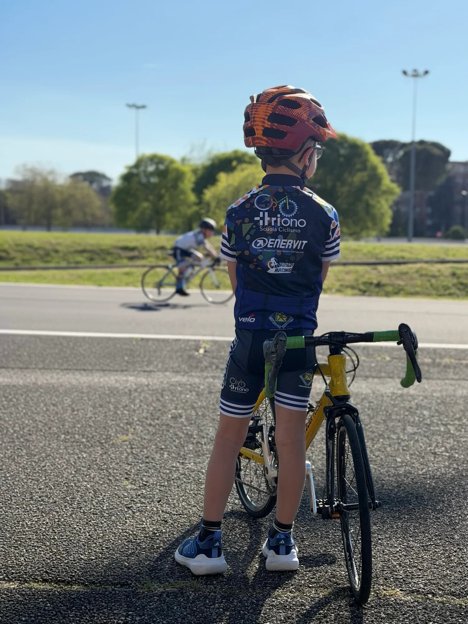 Allievo della Scuola Triono di spalle, fermo con la bici a bordo pista mentre un compagno pedala alle sue spalle