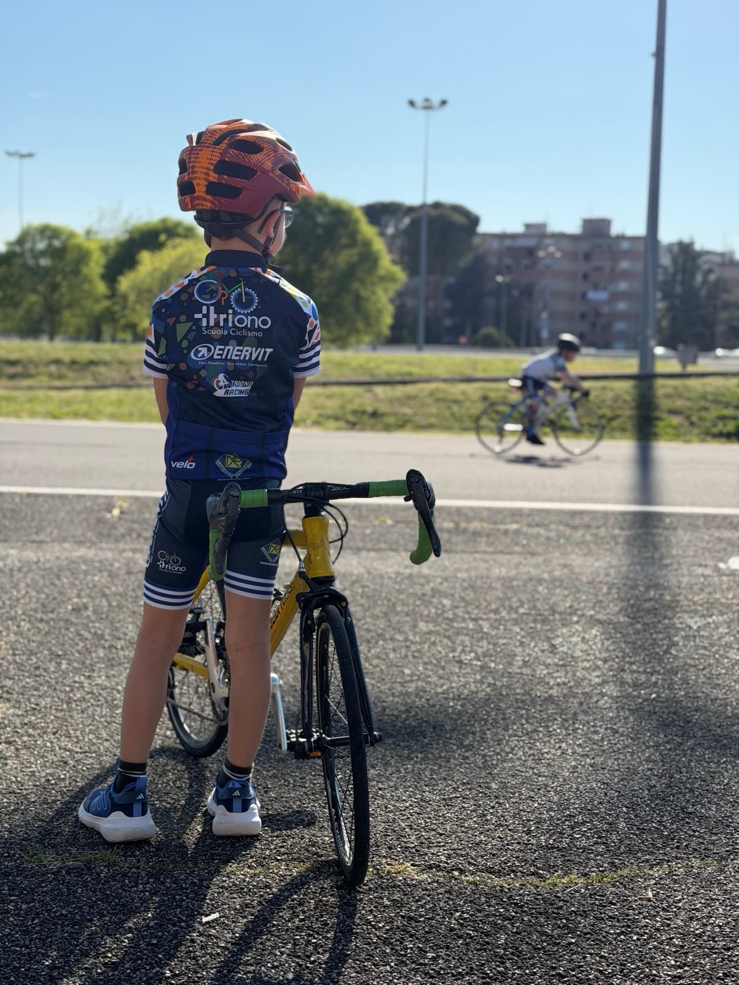 Bambino di spalle in divisa Triono osserva un compagno pedalare sulla pista del ciclodromo
