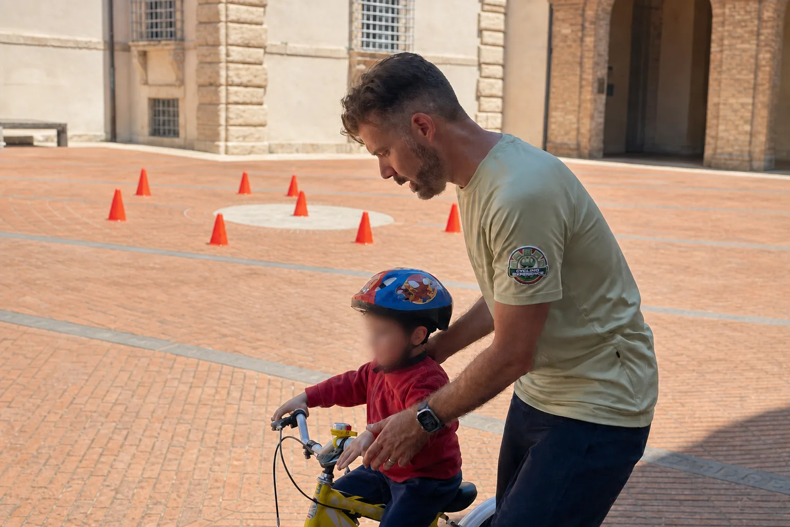Un maestro sostiene per le spalle un bambino in sella a una bici gialla, in una piazza pavimentata con alcuni coni arancioni disposti a terra