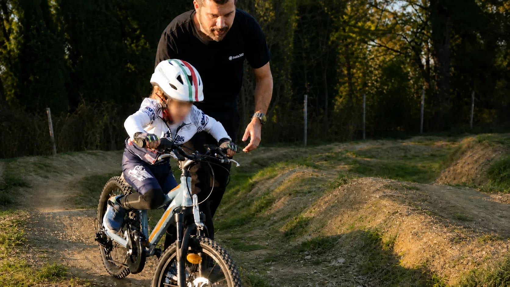 Un maestro cammina accanto a un bambino in mountain bike su un tracciato sterrato, con la luce bassa del tramonto