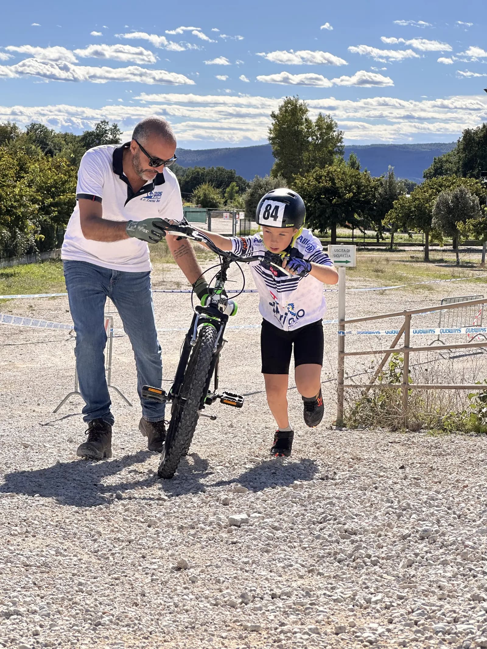 Un maestro tiene il manubrio della bici mentre un bambino con casco da gara la spinge su un percorso sterrato